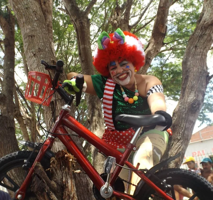 Tia Mila caracterizada de palhaça, sobre uma bicicleta, em evento comunitário